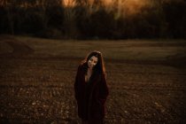 Donna gioiosa in cappotto caldo sorridente e guardando la fotocamera mentre cammina lungo la foresta autunnale — Foto stock