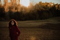 Радостная рыжая женщина в теплом пальто улыбается и смотрит в камеру во время прогулки по осеннему лесу — стоковое фото
