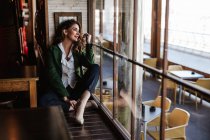 Зверху стомлена модна жінка сидить з схрещеними ногами і дивиться крізь вікно сучасного кафе — стокове фото