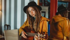 Молода стильна жінка з дредами в жовтому пальто і чорний капелюх сидять на старому дерев'яному столі назад до дзеркала і грають на гавайській гітарі укулеле в кімнаті з антикварними меблями — стокове фото