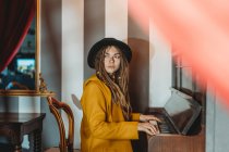 Seitenansicht einer ernsthaften Hipster-Frau mit Dreadlocks in gelbem Mantel und schwarzem Hut, die im retro gestylten Raum Klavier spielt — Stockfoto