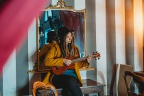Mulher elegante jovem com dreadlocks vestindo casaco amarelo e chapéu preto sentado na velha mesa de madeira de volta ao espelho e tocando ukulele guitarra havaiana — Fotografia de Stock