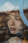 Pensive sad young female with dreadlocks leaning against window glass and looking away — Stock Photo