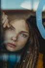 Pensive sad young female with dreadlocks leaning against window glass and looking away — Stock Photo