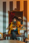 Young stylish woman with dreadlocks wearing yellow coat and black hat sitting on old wooden table back to mirror and playing Hawaiian guitar ukulele — Stock Photo