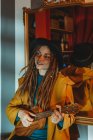 Молодая стильная женщина с дредами в желтом пальто и черной шляпе сидит на старом деревянном столе к зеркалу и играет на гавайской гитаре укулеле — стоковое фото