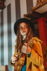 Young stylish woman with dreadlocks wearing yellow coat and black hat sitting on old wooden table back to mirror and playing Hawaiian guitar ukulele — Stock Photo