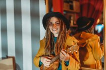 Молода стильна весела жінка з дредами в жовтому пальто і чорний капелюх сидять на старому дерев'яному столику назад до дзеркала і грають на гавайській гітарі укулеле. — стокове фото