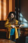 Hipster-Millennial-Frau mit Dreadlocks in gelbem Mantel und schwarzem Hut spielt hawaiianische Gitarrenukulele, während sie im dunklen Vintage-Raum steht — Stockfoto