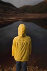 Visão traseira da pessoa em capa de chuva amarela com capuz cobrindo a cabeça em pé perto do lago no dia nublado — Fotografia de Stock