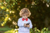 Маленький кудрявый мальчик в рубашке и красной бабочке с помощью мобильного телефона с зелеными растениями на размытом фоне — стоковое фото