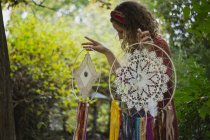Frau in Freizeitkleidung zeigt handgefertigte Traumfänger mit langen Fäden beim Betrachten von Amuletten, die Zeit an der frischen Luft verbringen — Stockfoto