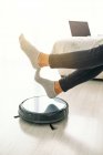 Side view of crop female in jeans and socks sitting on white sofa in room with laminate floor and putting feet on robotic vacuum cleaner — Stock Photo