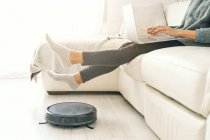 Side view of crop female in jeans and socks sitting on white sofa working on computer in room with laminate floor and putting feet on robotic vacuum cleaner — Stock Photo