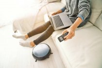 From above side view of crop female in casual clothes using remote control for robotic vacuum cleaner while sitting on sofa and enjoying free time browsing internet on laptop — Stock Photo