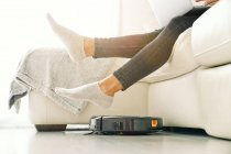 Seitenansicht einer Frau in Jeans und Socken, die auf einem weißen Sofa im Zimmer mit Laminatboden sitzt und ihre Füße auf einen Staubsaugerroboter setzt — Stockfoto