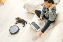 From above woman sitting on sofa in light room with cute puppy lying down next round black robotic vacuum cleaner — Stock Photo