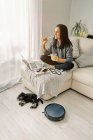 Женщина сидит на диване в светлой комнате, работает за компьютером и пьет холодный напиток с милым щенком лежащим рядом круглый черный роботизированный пылесос — стоковое фото