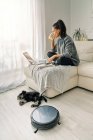 Donna seduta sul divano in una stanza luminosa che lavora su un computer e beve bevande fredde con cucciolo carino sdraiato accanto all'aspirapolvere robotico nero rotondo — Foto stock