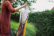 Cropped anonymous female in casual wear showing handmade dreamcatchers with long threads looking at amulet spending time in fresh air — Stock Photo