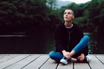 Female hipster with short hairstyle in casual clothing sitting with crossed legs on wooden pier of pond with green trees on blurred background — Stock Photo