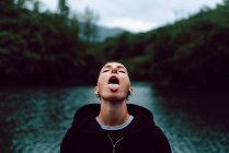 Kurzhaarige Frau mit Piercing trägt schwarzen Kapuzenpulli und schaut auf, während sie Regentropfen mit Zunge in der Nähe von grünem Wald und Teich fängt — Stockfoto