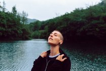 Woman with short hairstyle and piercing in casual clothing facing head up with closed eyes to sky with pond among green plants on blurred background — Stock Photo