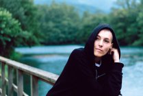 Frau in schwarzem Kapuzenpulli lehnt an Holzzaun der Brücke und schaut weg mit türkisfarbenem Teich und grünen Pflanzen auf verschwommenem Hintergrund — Stockfoto
