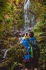 Обратный вид женщины с профессиональной камерой и рюкзаком, смотрящей в сторону, стоя у водопада в лесу в летний день — стоковое фото