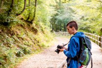 Серйозний юнацький жінка-пішохід, насолоджується відпусткою і фотографується на професійній камері, стоячи на дерев'яній доріжці в лісі — стокове фото