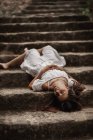 Dall'alto di tenera affascinante giovane donna in abito bianco sdraiato vulnerabilmente sulle scale nel parco autunnale guardando altrove — Foto stock