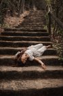 From above of tender charming young woman in white dress lying down vulnerably on stairs in autumnal park looking away — Stock Photo