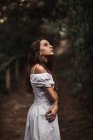 Side view of tender gorgeous young woman in white dress embracing shoulders standing in calm park and looking away — Stock Photo