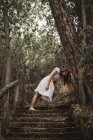 Dal basso di misteriosa donna calma in abito bianco appoggiata con la testa sul tronco d'albero in piedi sulle scale in un tranquillo parco autunnale — Foto stock