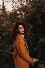 Жінка з темним кучерявим волоссям у в'язаному джемпері та пальто, стоячи в парку, кладе руку на металеві перила, дивлячись на камеру — стокове фото