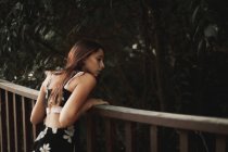 Vue arrière de la femme tendre en robe élégante appuyée sur une clôture en bois et regardant vers le bas dans un parc paisible — Photo de stock