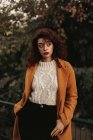 Donna con i capelli ricci scuri indossa maglione a maglia e cappotto in piedi nel parco mettendo mano su ringhiera metallica mentre guarda la fotocamera — Foto stock