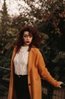 Woman with dark curly hair wearing knitted jumper and overcoat standing in park putting hand on metal railing while looking at camera — Stock Photo