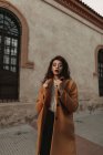 Donna in maglia calda maglione e cappotto con le braccia alzate in piedi accanto all'edificio mentre guarda la fotocamera con sfida — Foto stock