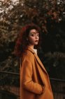 Donna con i capelli ricci scuri indossa maglione a maglia e cappotto in piedi nel parco mettendo mano su ringhiera metallica mentre guarda la fotocamera — Foto stock