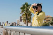 Giovane fotografo di sesso maschile ispirato in felpa casual gialla che parla foto sulla macchina fotografica in piedi sulla banchina della città alla luce del sole — Foto stock