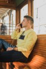 Vue latérale du jeune homme en vêtements décontractés parlant sur un téléphone portable assis sur un banc en bois dans un vieux train — Photo de stock