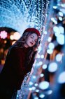 Elegante donna in abbigliamento alla moda e cappello vicino al muro con luci nel periodo natalizio — Foto stock