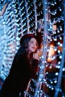Femme élégante en vêtements à la mode et chapeau près du mur avec des lumières pendant la période de Noël — Photo de stock