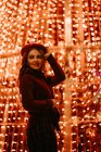 Elegante Frau in trendiger Kleidung und Hut an der Wand mit Lichtern in der Weihnachtszeit — Stockfoto