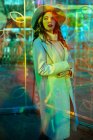 Стильна молода жінка в білому пальто дивиться у світлі неонових знаків на міській вулиці — стокове фото