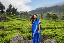 Vista lateral da mulher asiática em sari azul em pé na rocha olhando para a câmera no meio de campos de chá em Haputale, no Sri Lanka — Fotografia de Stock