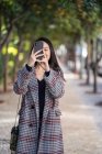 Азиатка в клетчатой куртке с сумочкой, фокусирующаяся на экране и снимающая со смартфона — стоковое фото