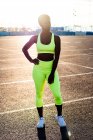 Mujer afroamericana enfocada en serio en vibrante ropa deportiva amarilla mirando hacia otro lado y contemplando mientras está de pie con la mano en la cadera en los rayos de sol solos en la calle contra el medio ambiente urbano - foto de stock