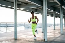 African American adult sportswoman in vibrant green activewear focusing and running alone along waterfront among metal columns under roof — Stock Photo